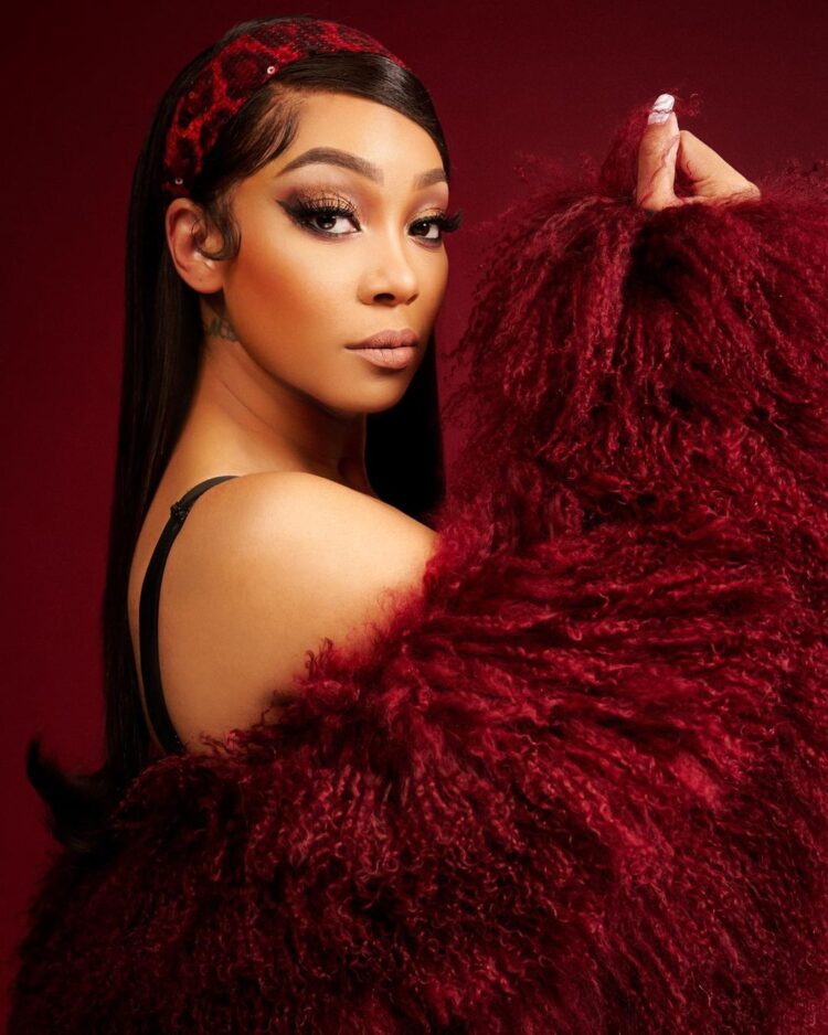 R&B singer Monica holding up a plush red coat to the side of her face, posing against a red background. She is wearing a matching red headband and elaborate eye makeup, looking directly at the camera.