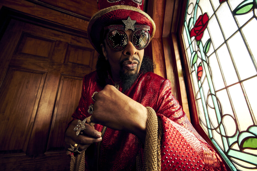 A photo of Bootsy Collins wearing hat and sunglasses poses in front of colorful stained glass window.