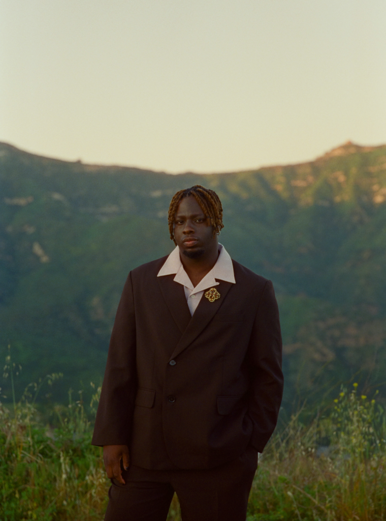 Blk Odyssy wearing a dark suit adorned with a distinctive brooch stands in a natural setting with lush, green hills in the background, projecting a sense of serene contemplation.