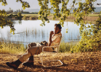 The image features singer Leon Bridges seated in a relaxed pose on a folding chair beside a serene lake, surrounded by grassy fields and trees under clear skies. He is wearing a casual, patterned short-sleeved shirt and light-colored pants, with one hand touching his chin thoughtfully. In the upper left corner, the text "Leon" appears in large letters. The lower right corner contains the Columbia brand logo.