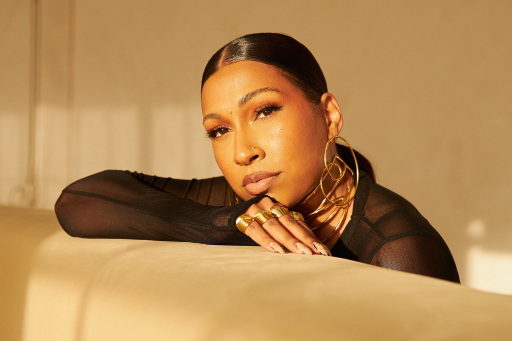 Portrait of a Melanie Fiona resting her head on her arms on a beige couch, lit by warm sunlight. She is wearing a black mesh top and large gold hoop earrings.