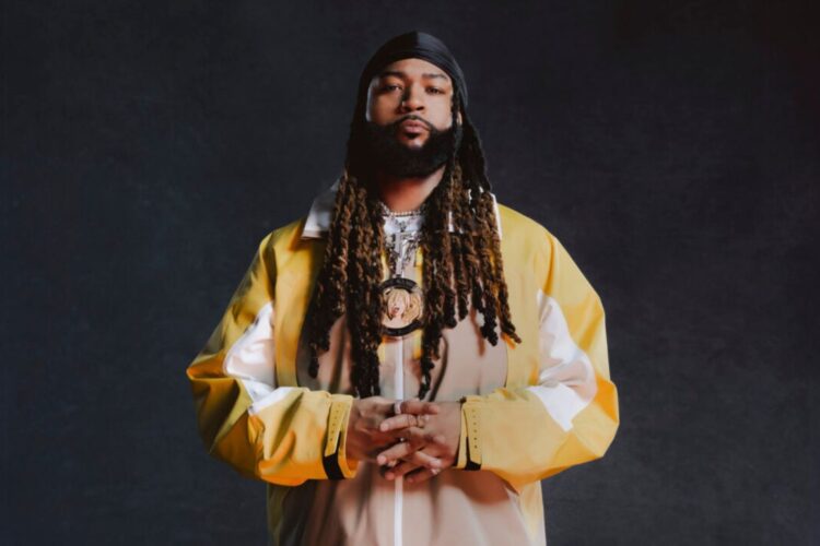 In image of singer PartyNextDoor wearing a yellow, white and tan jacket against a dark grey backdrop.