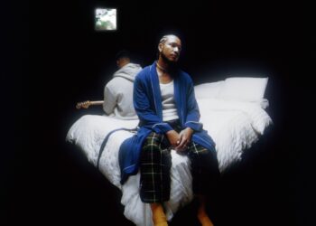 R&B duo Bathe's Inside Voice(s) album cover: Devin Hobdy sits on a bed in a blue robe and pajama pants, while Corey Smith-West, in a gray hoodie, holds a guitar. The bed is dressed with white comforters in a dark room, illuminated solely by light streaming through a small window.