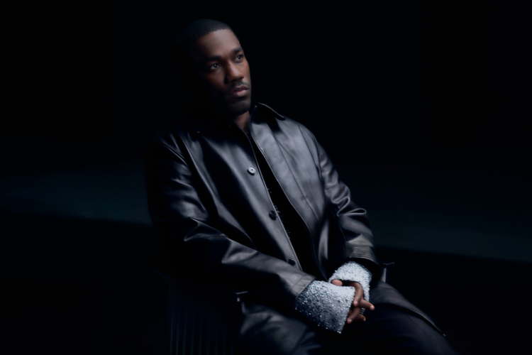 R&B singer Giveon, wearing a leather black jacket, seated in front of a black backdrop.